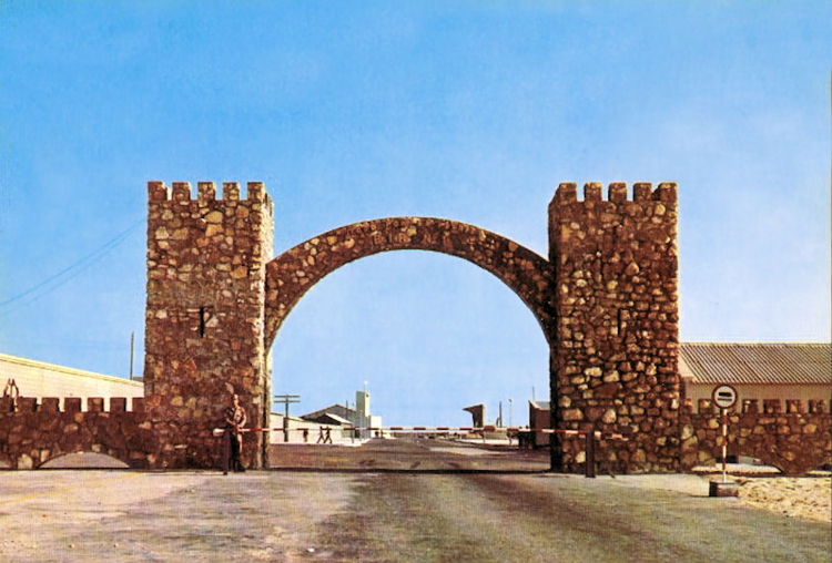 03-01.- El arco de entrada a El BIR, el arco más famoso del Sáhara. Dicen que se edificó para poder colgar en él lo que había que colgar a la entrada. Entre este arco, el océano y el Campo de Margaritas, se sufría una dura instrucción. Postal: 1975